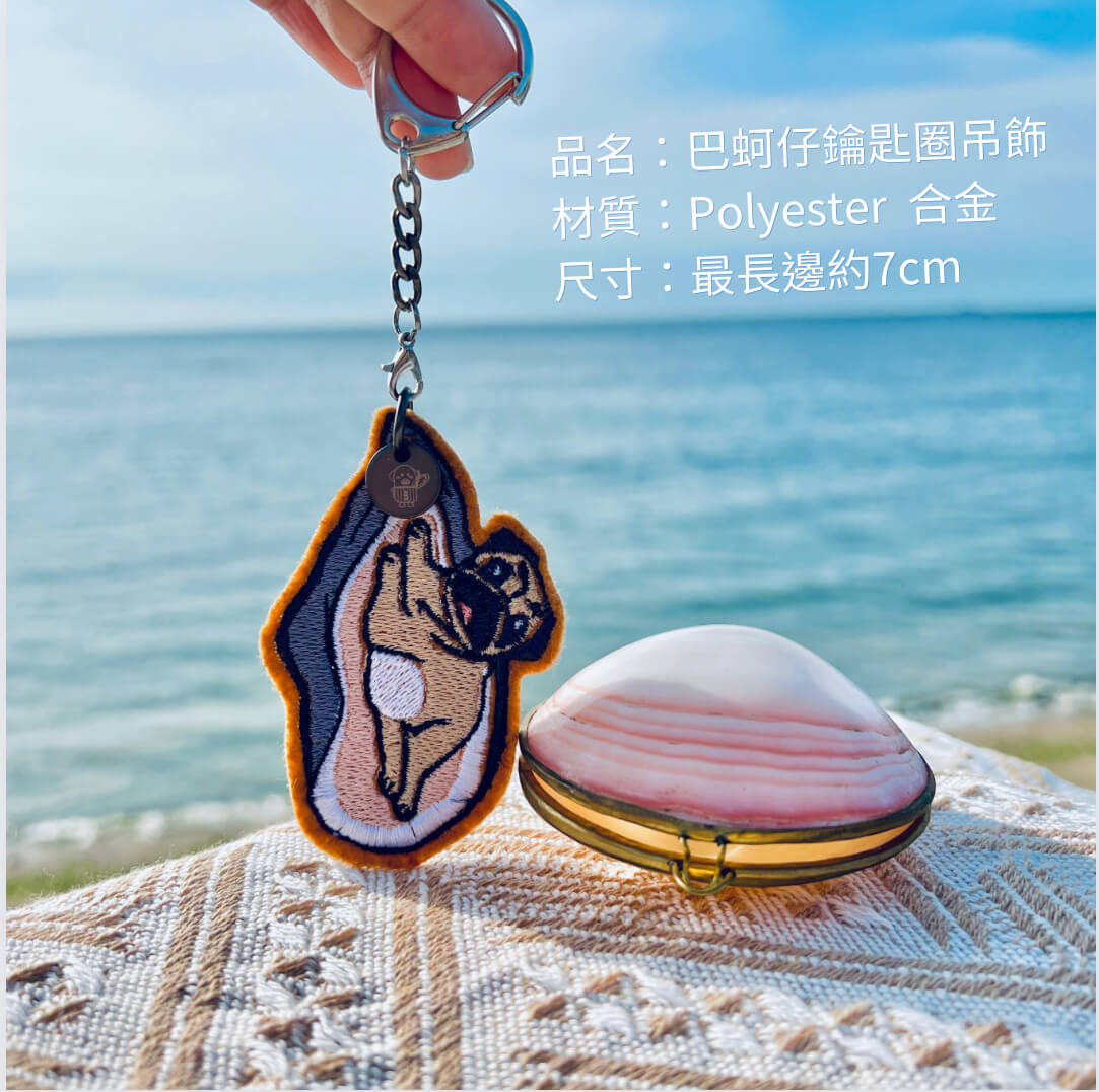 雙面刺繡巴哥犬蚵仔鑰匙圈吊飾 3 手中拿著巴哥犬變身蚵仔刺繡鑰匙圈,旁邊放置一個粉色條紋貝殼